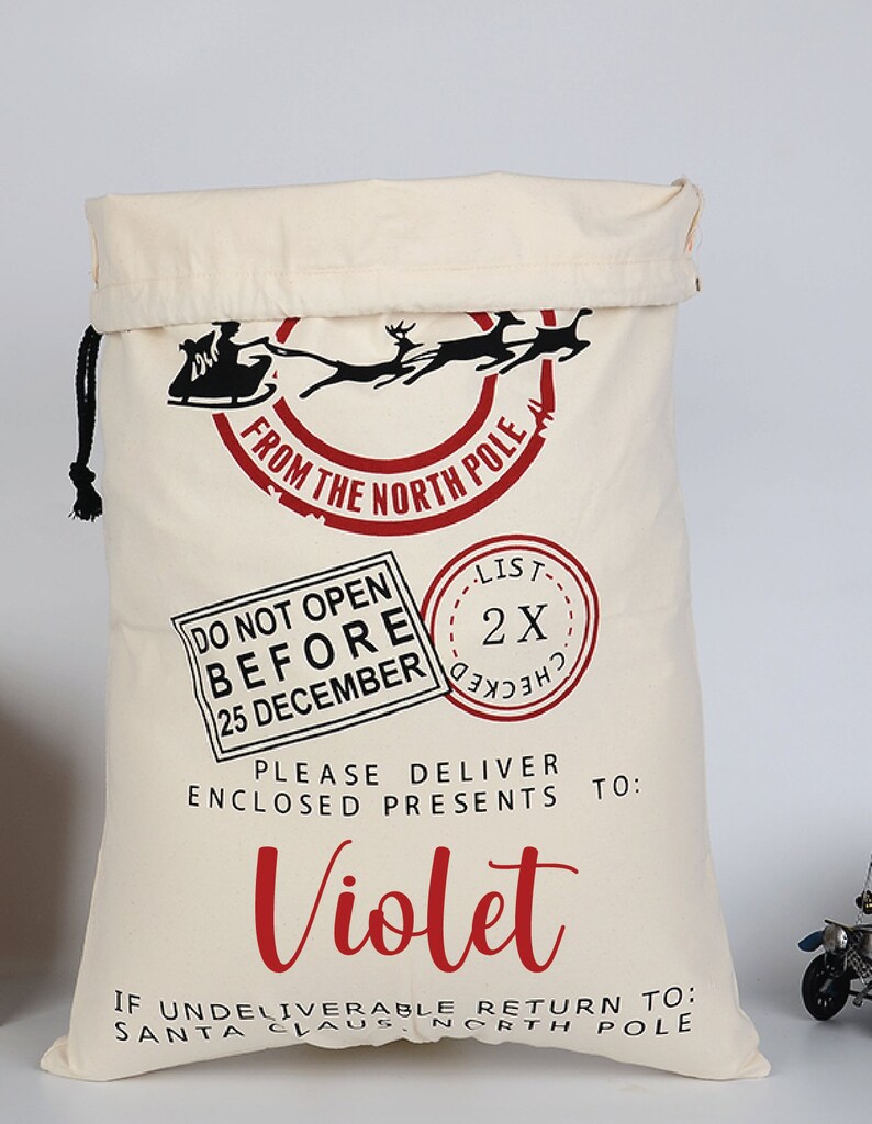 Pu&ograve; includere: Un sacco di Babbo Natale in tela bianca con un design rosso e nero. Il sacco ha una chiusura a coulisse e dice "From the North Pole" in un cerchio rosso. Dice anche "Do Not Open Before 25 December" e "Please Deliver Enclosed Presents To: Violet".