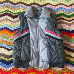 May include: A gray and light gray vintage vest with a high collar and a full zipper. The vest has a black, pink, and light blue stripe design on the sides. The vest has two pockets and the brand name "GINGO SCENE" is visible on the collar.