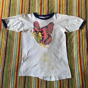 May include: White ringer t-shirt with a colorful butterfly graphic. The butterfly has pink, blue, and yellow details. The shirt has a navy blue collar and sleeve trim. The shirt is vintage and shows signs of wear.