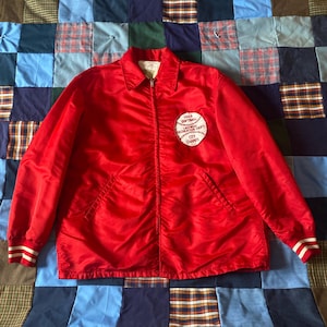 May include: A bright red vintage jacket with a white patch that reads "1968 Softball League Dedication Dept City Champs." The jacket has a zipper closure, a collar, and white stripes on the cuffs. The jacket is made of a shiny material.