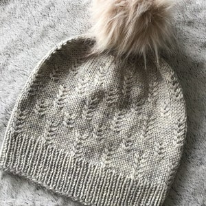 May include: A knitted beanie hat with a textured pattern and a fluffy pom pom on top. The hat is a light beige color.