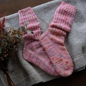 Op de afbeelding: Een paar roze en veelkleurige gebreide sokken met geribbelde manchetten, rustend op een grijze gebreide deken. De sokken hebben een gespikkeld patroon en zijn omgeslagen. Een klein boeket droogbloemen staat op de achtergrond.