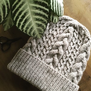 May include: A gray knitted hat with a cable knit pattern. The hat has a folded brim.