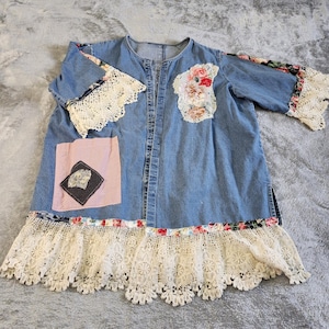 May include: A light blue denim tunic with floral and lace accents. The tunic features a button-up front, short sleeves with lace trim, and a large lace hem. A pink patch with a black and white design is on the front.