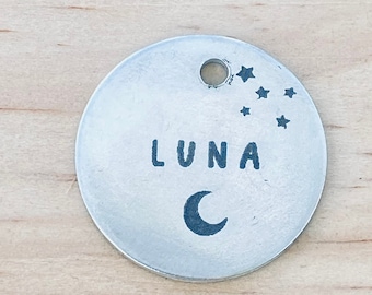 Könnte beinhalten: Ein runder Anhänger aus Silber mit dem eingravierten Namen "LUNA". Der Anhänger zeigt außerdem einen Halbmond und eine Gruppe von Sternen.