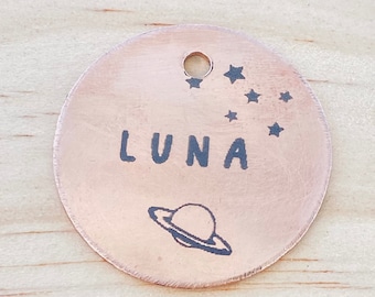 Könnte beinhalten: Ein runder Hundemarken aus Kupfer mit der Gravur "LUNA". Die Marke zeigt außerdem einen kleinen Planeten und eine Sternenkonstellation.