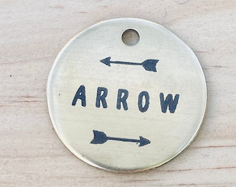 Könnte beinhalten: Ein runder, goldfarbener Metallschild mit dem Wort "ARROW" in schwarzen Lettern eingraviert. Zwei schwarze Pfeile zeigen in entgegengesetzte Richtungen über und unter dem Wort.