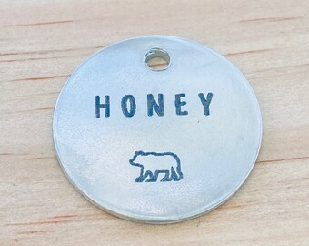 Könnte beinhalten: Runder silberner Haustieranhänger mit dem Wort "HONEY" in Großbuchstaben und einer kleinen Bärensilhouette.
