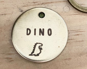 Può includere: Un'etichetta rotonda in ottone con una silhouette di dinosauro e la parola "DINO" incisa su di essa. L'etichetta ha una piccola pietra preziosa verde nella parte superiore.