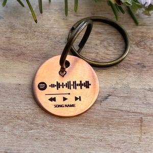 Puede incluir: Un llavero redondo de metal color cobre con una interfaz de reproductor de música grabada con láser. El diseño incluye un icono de corazón, un gráfico de onda sonora, símbolos de control de reproducción y el texto "SONG NAME" en letra sans-serif.