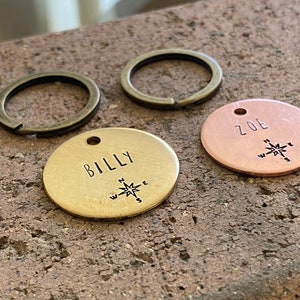 Könnte beinhalten: Zwei personalisierte Hundemarken mit Kompass-Designs. Eine Marke ist aus Messing mit dem eingravierten Namen "BILLY". Die andere Marke ist aus Kupfer mit dem eingravierten Namen "ZOE".