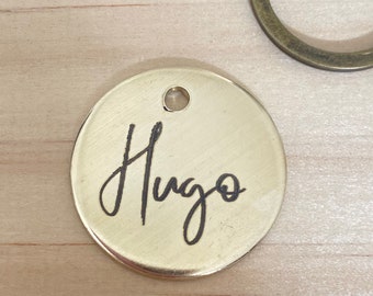 May include: A round metal tag in gold with a hole at the top and the name "Hugo" engraved in black cursive.