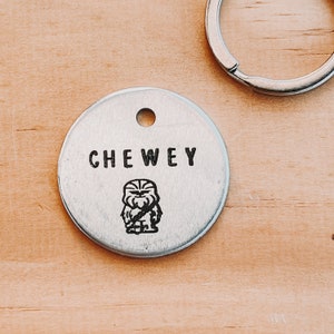 Op de afbeelding: Een zilveren metalen hondenpenning met de naam "CHEWEY" erop gegraveerd. Onder de naam staat een kleine zwart-witte illustratie van een personage uit Star Wars.