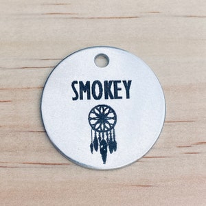 以下が含まれることがあります： 黒い文字で「SMOKEY」と刻まれた銀色の丸い犬用タグ。名前の下には黒いドリームキャッチャーのデザインがあります。