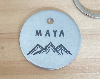 May include: Silver round tag with the name "MAYA" engraved in black lettering and a mountain range design.