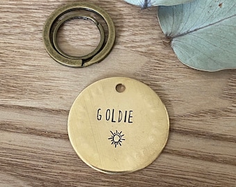 Könnte beinhalten: Ein rundes Messing-Schild mit dem eingravierten Namen "Goldie" und einem kleinen Sonnensymbol.