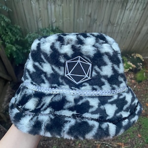 May include: A black and white faux fur bucket hat featuring a central embroidered patch of a twenty-sided die. The hat is accented with a silver braided trim above the brim and a row of black sequins along the edge of the brim.