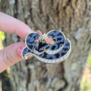 Op de afbeelding: Een gouden emaille pin met een zwart-witte slang met een bloemenkroon. De slang is opgerold met zijn kop naar de kijker gericht. De bloemen zijn roze en wit.