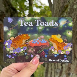 Könnte beinhalten: Zwei goldene Emaille-Pins mit braunen Fröschen mit gelben Akzenten, die in einem Bett aus lila und weißen Blumen sitzen. Der Text "Tea Toads" und "Heartel Design" ist auf der Karte sichtbar.