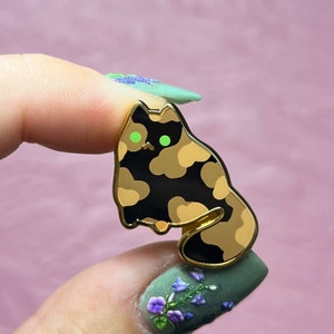 Könnte beinhalten: Goldfarbene Emaille-Anstecknadel mit einem Cartoon-Katzenmotiv mit schwarzen und braunen Tarnflecken und grünen Augen.