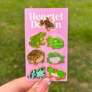 Puede incluir: Un conjunto de siete pines de esmalte de rana en una tarjeta rosa con el texto "Heartel Design". Los pines presentan diferentes especies de ranas en varios colores, incluidos el verde, el marrón y el azul.