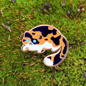 May include: A gold-colored enamel pin featuring a cartoon calico cat with black, orange, and white markings. The cat has blue eyes and is curled up in a sleeping position.