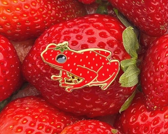 May include: A red enamel pin of a frog wearing a crown of leaves. The frog is sitting on a bed of red strawberries.