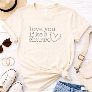 May include: Cream-colored t-shirt with the phrase "love you like a churro" in gray text and a heart graphic. The shirt is knotted at the waist. Accessories include sunglasses, a straw hat, and white sneakers.