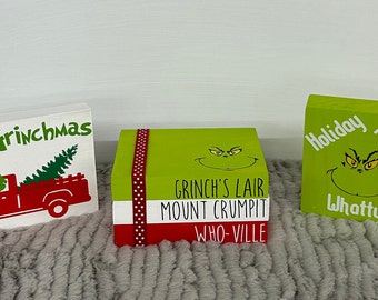 May include: Three decorative wooden blocks with a Christmas theme. One block features a red truck carrying a Christmas tree with the text "Merry Grinchmas." Another block has the Grinch's face and the text "Grinch's Lair, Mount Crumpit, Who-Ville." The third block has the Grinch's face and the text "Holiday Hoobie Whatty?"