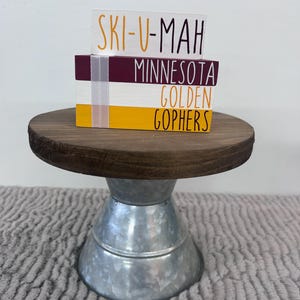 May include: A decorative wooden stand with a galvanized metal base. The stand holds three stacked wooden blocks with the words "SKI-U-MAH", "MINNESOTA", and "GOLDEN GOPHERS" in black and gold lettering. A white ribbon is wrapped around the blocks.