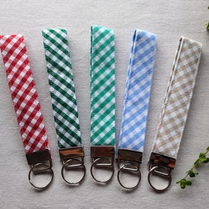 May include: Five fabric wristlet keychains featuring a diagonal gingham pattern in red, dark green, bright green, light blue, and beige. Each strap is attached to a silver-toned metal hardware clamp with a circular split ring for holding keys.