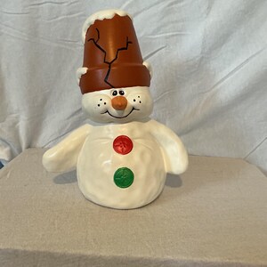 Könnte beinhalten: Keramik-Schneemannfigur mit braunem Eimerhut und Karottennase. Der Schneemann hat einen weißen Körper mit roten und grünen Knöpfen. Der Hut hat ein Rissmuster. Eine festliche Dekoration für das Zuhause.