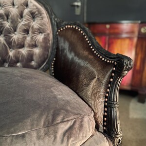 May include: A gray velvet tufted sofa with a brown cowhide back and black wooden frame. The sofa has a decorative carved design on the frame.