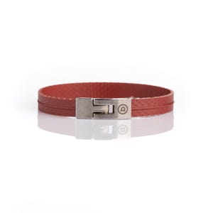 Könnte beinhalten: Ein rotes Armband mit strukturierter Oberfläche und einer horizontalen Linie in der Mitte. Das Armband verfügt über einen rechteckigen Verschluss aus silberfarbenem Metall mit einem kleinen kreisförmigen Logo, in das der Buchstabe a eingraviert ist.