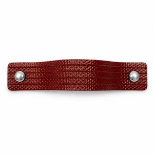 Peut inclure: Une poignée de meuble rouge foncé avec une surface texturée rappelant le cuir et des lignes horizontales rainurées. La poignée présente une forme arquée et est fixée aux deux extrémités par des rivets métalliques circulaires argentés.