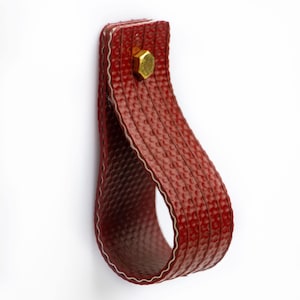 Könnte beinhalten: Ein roter Leder-Schlaufen-Griff mit einer Messing-Schraube. Der Griff ist strukturiert und hat eine leicht abgerundete Form.