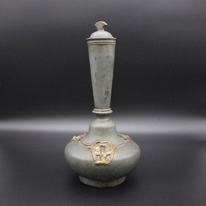 May include: A gray ceramic vase with a gold lid and decorative accents. The vase has a round body and a tall, slender neck.