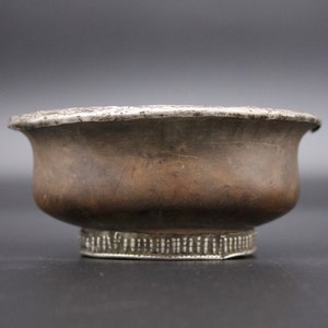 Puede incluir: Un cuenco de plata con un fondo ligeramente redondeado y un borde decorativo. El cuenco tiene un aspecto desgastado y envejecido.