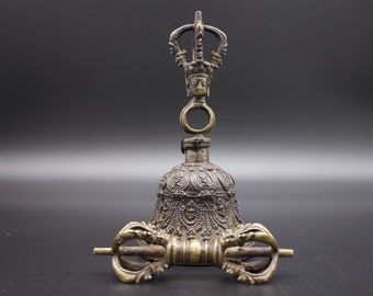 May include: A silver-colored bell with intricate carvings and a small figure on top. The bell is attached to a handle with a ring.