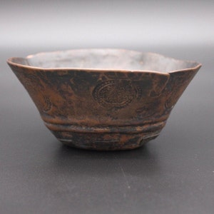 May include: A small, copper bowl with a hammered finish and a decorative pattern of circles. The bowl has a slightly irregular shape and shows signs of age and wear.
