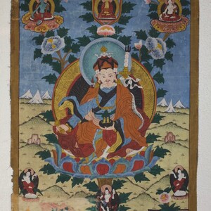 Puede incluir: Una pintura budista tibetana colorida que representa una deidad sentada en un trono de loto. La deidad est&aacute; rodeada de figuras m&aacute;s peque&ntilde;as y motivos florales. La pintura est&aacute; enmarcada en oro y tiene un fondo azul.