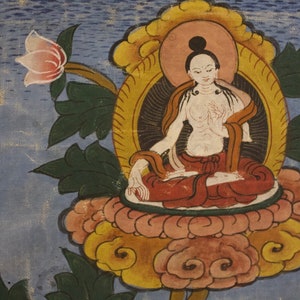 Puede incluir: Una pintura de una deidad sentada en posici&oacute;n de loto sobre una flor de loto. La deidad lleva una t&uacute;nica roja y tiene un cuerpo blanco. El fondo es azul y tiene un dise&ntilde;o floral.