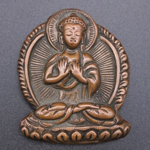 May include: A copper-colored Buddha statue pendant with a detailed design. The Buddha is seated in a meditative pose with hands clasped in front of the chest. The pendant is surrounded by a decorative halo.