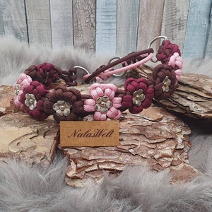 Könnte beinhalten: Ein braunes und pinkfarbenes Paracord-Hundehalsband mit silbernen Blumenakzenten. Das Halsband ist aus einem Paracord-Seil gefertigt und hat eine Metallschnalle. Das Halsband liegt auf einer Holzoberfläche mit einem braunen Etikett, auf dem "NalasWell" steht.