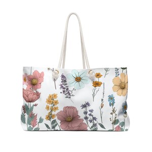 May include: A white canvas tote bag with a floral print featuring pink, yellow, blue, and green flowers. The bag has white rope handles.