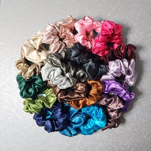 Pu&ograve; includere: Una collezione di 20 scrunchies di raso colorati in varie tonalit&agrave; di verde, blu, marrone, rosa, viola e nero. Gli scrunchies sono disposti in un cerchio su una superficie grigia.