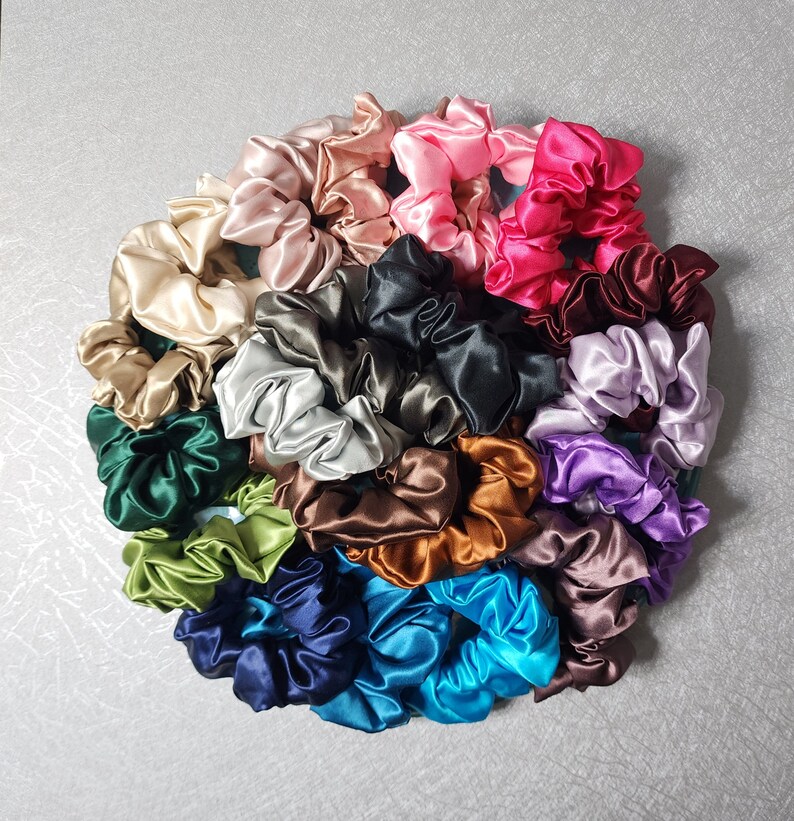 Pu&ograve; includere: Una collezione di 20 scrunchies di raso colorati in varie tonalit&agrave; di verde, blu, marrone, rosa, viola e nero. Gli scrunchies sono disposti in un cerchio su una superficie grigia.