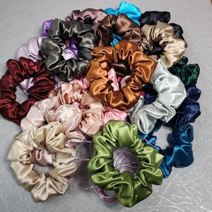 Pu&ograve; includere: Una collezione di scrunchies di raso colorati in varie tonalit&agrave; di blu, verde, viola, marrone, rosso e bianco. Gli scrunchies sono disposti in una pila, mostrando la loro texture morbida e setosa.