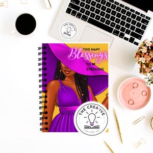 Può includere: Un taccuino a spirale con un'illustrazione di una donna con un vestito e un cappello viola. Il testo "TOO MANY Blessings TO BE STRESSING" è visibile. Il taccuino è circondato da forniture per ufficio e un laptop.