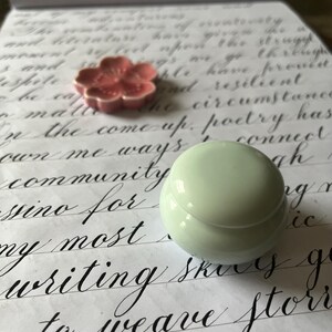 May include: A close-up of a white ceramic container with a lid, sitting on a piece of paper with handwritten text. The text includes the words "community", "poetry", and "writing".